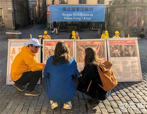 '图6:两位来自隆德大学的年轻人一直是蹲在展板前了解真相,学员(左一)看到此景就蹲在他们身旁耐心的讲真相。'