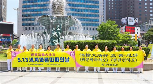 图1:5月13日,部分首尔西部和中部的法轮功学员们在首尔市中心新世界百货店附近的大型喷泉前举行了集体炼功、派发传单等洪法活动。图为他们在活动前,首先恭祝慈悲伟大的师尊华诞。